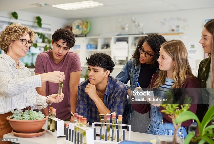 Mature science teacher showing sample in test tube to a group of multiethnic students during classroom activity. High school guys and girls gathered around table to observe biology experiment involving plant specimen. Group of young college classmates working on science project at school.